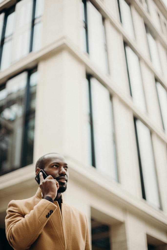 Confident man in a trench coat making a call outdoors in front of a modern building.