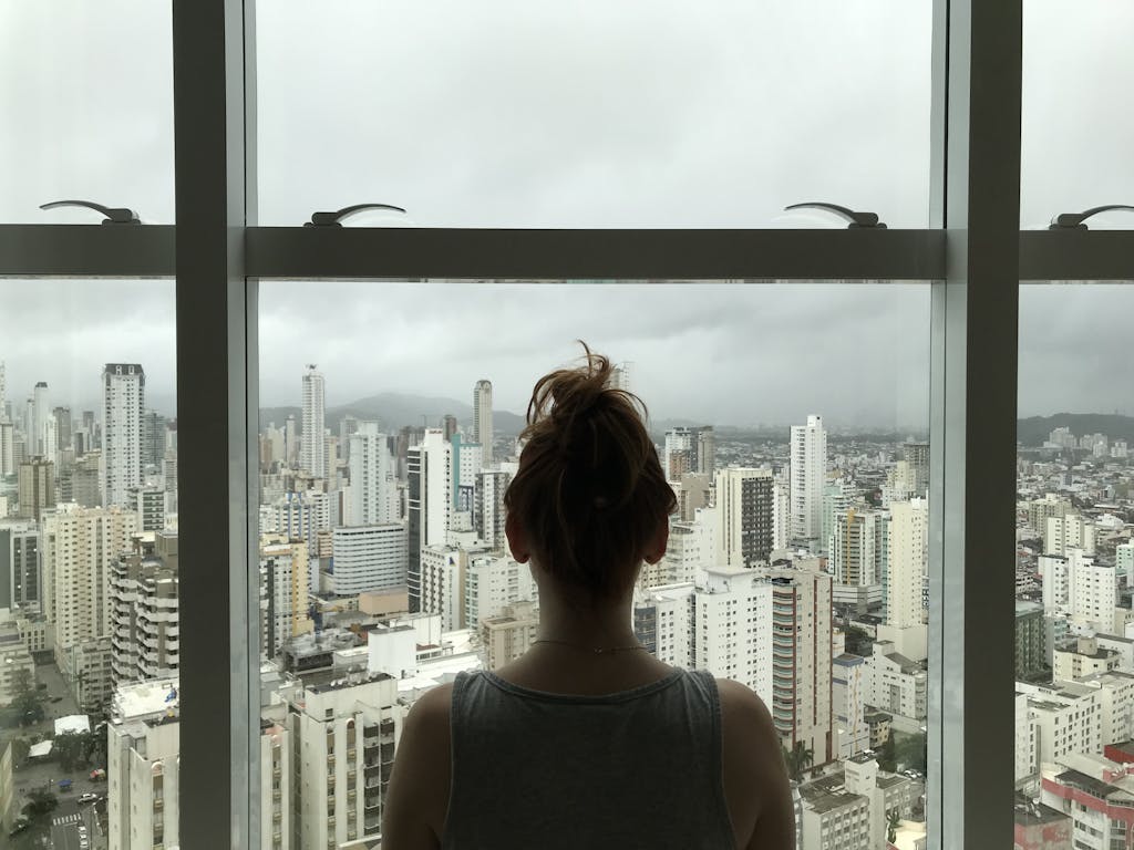 A woman gazing at a cloudy city skyline through a large window, capturing urban life.